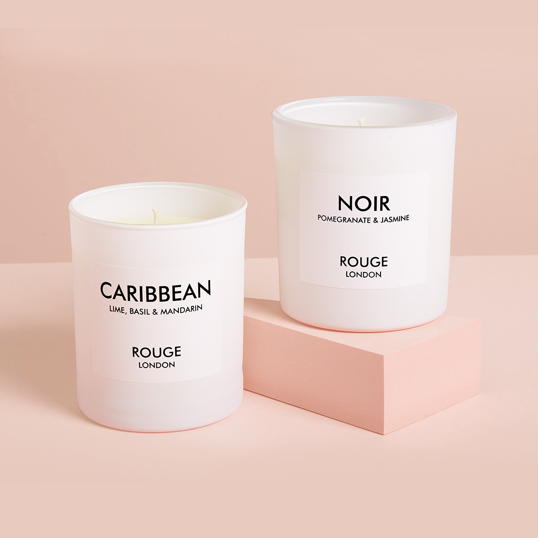 Rouge London Two white candles, one labeled 'Caribbean' and the other 'Noir', placed on a pink surface against a pink background.