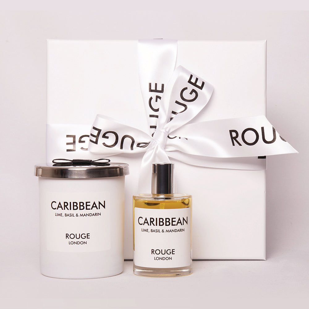 A gift set by Rouge London including a white candle in a container and a spray bottle, both labeled 'Caribbean - Lime, Basil & Mandarin'. The items are presented in a white box with a black ribbon.