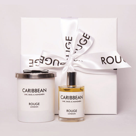 A gift set by Rouge London including a white candle in a container and a spray bottle, both labeled 'Caribbean - Lime, Basil & Mandarin'. The items are presented in a white box with a black ribbon.