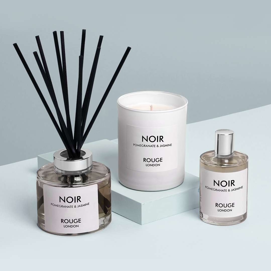 A luxury gift collection set including a white candle in a jar, a reed diffuser with black sticks, and a small spray bottle, all labeled 'NOIR' and 'ROUGE LONDON'. Presented on a two-tone background, light blue and white.